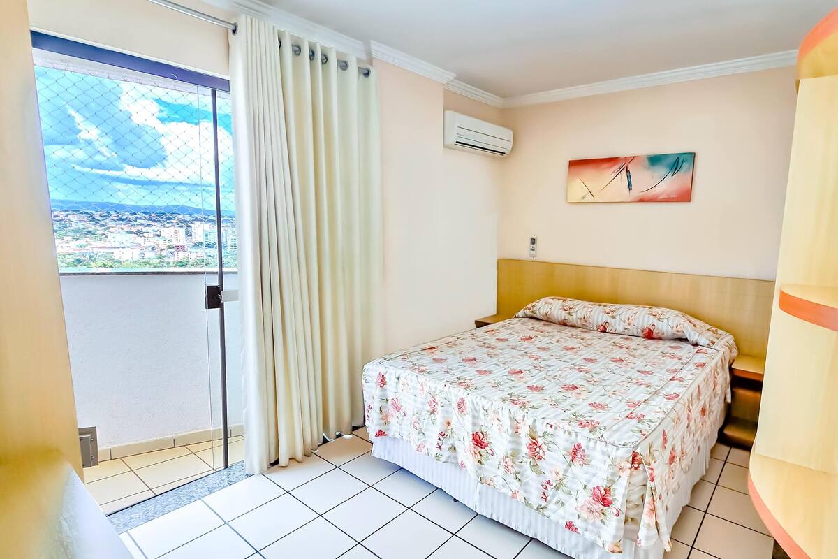 A hotel room with a bed, a view of a cityscape through a window, an air conditioning unit, and a painting on the wall.