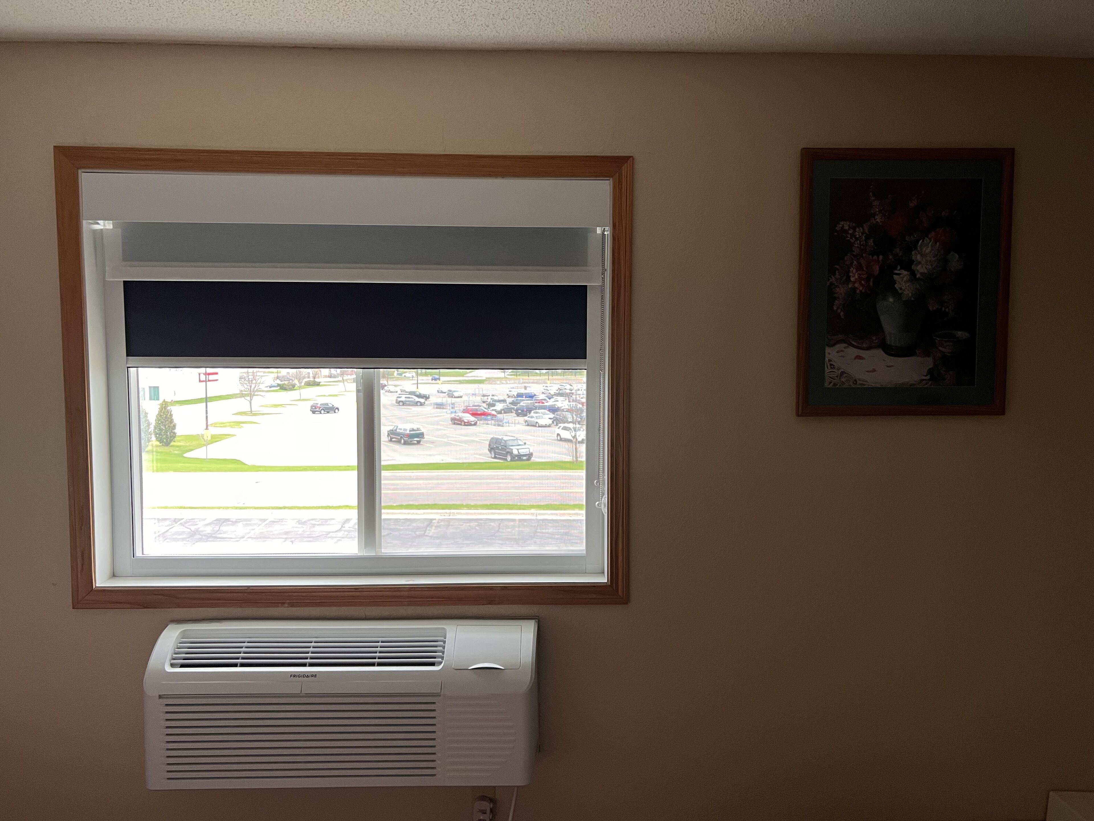 A room with a window overlooking a parking lot, a wall-mounted air conditioner, and a framed picture.
