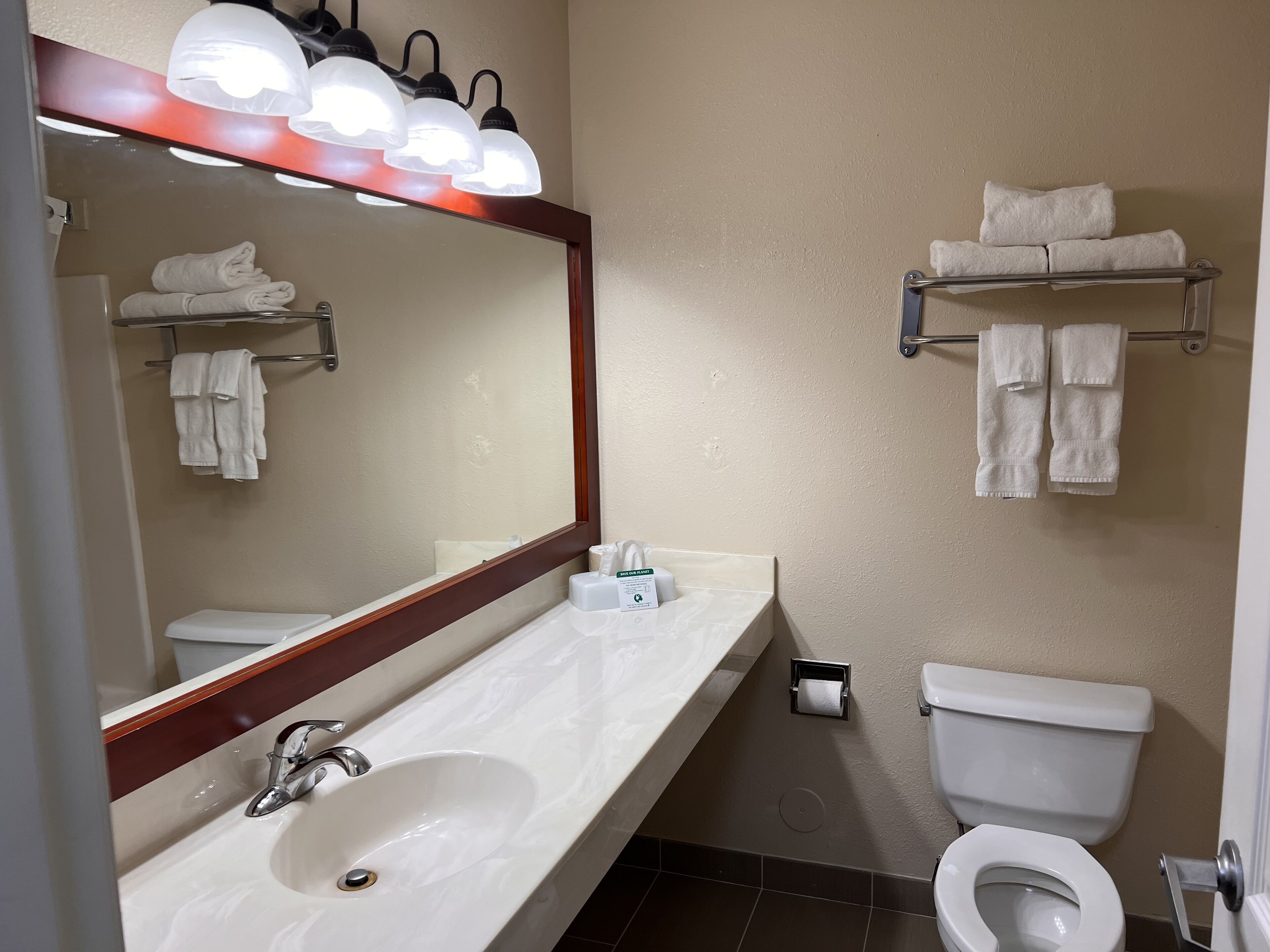 Un baño con lavamanos blanco, espejo y retrete. Hay toallas colgadas en un perchero y un dispensador de toallas.