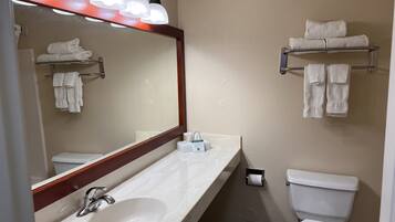 Un baño con lavamanos blanco, espejo y retrete. Hay toallas colgadas en un perchero y un dispensador de toallas.