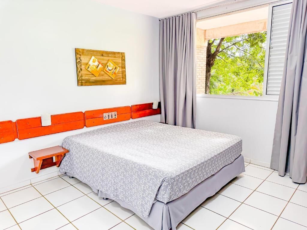 Ein Schlafzimmer mit einem Bett, einem kleinen Holzschemel, einer Wandsteckdose, einem Fenster mit Vorhängen und einem gerahmten Bild an der Wand.