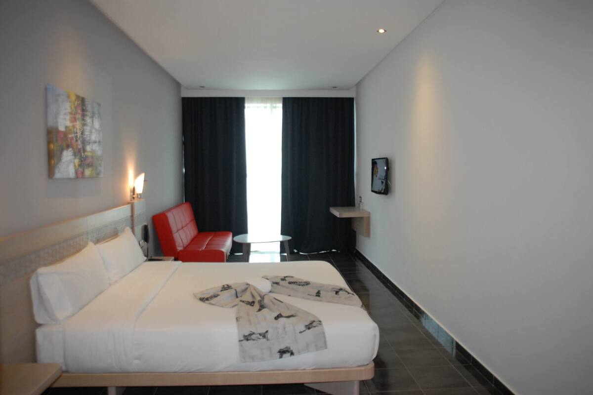 A hotel room with a large bed, a red chair, a small table, and a view of the outdoors through the window.
