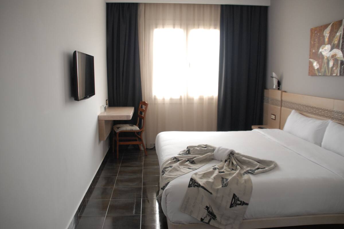 A hotel room with a bed, a desk, a chair, a TV, and a window with curtains.