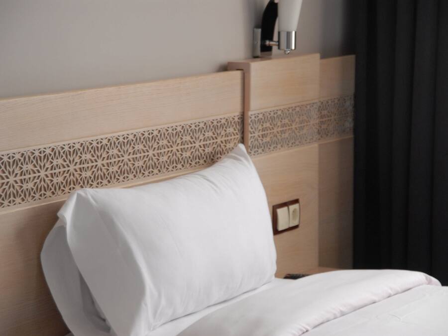 Ein ordentlich bezogenes Bett mit einem hölzernen Kopfteil mit geometrischem Muster, einem weißen Kissen und einem Nachttisch mit Lampe.
