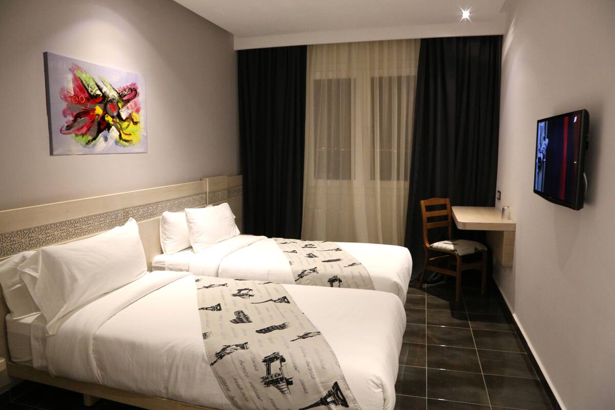 A hotel room with two beds, a desk, a chair, a TV, and a painting on the wall.