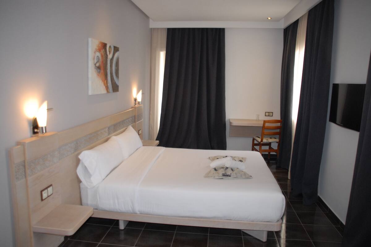 A hotel room with a bed, bedside table, lamp, artwork, and a desk with a chair.