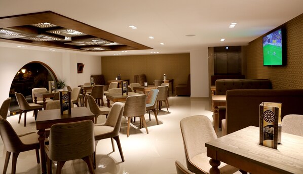 A modern dining area with wooden tables and chairs, recessed ceiling lighting, and a flat-screen TV mounted on the wall.