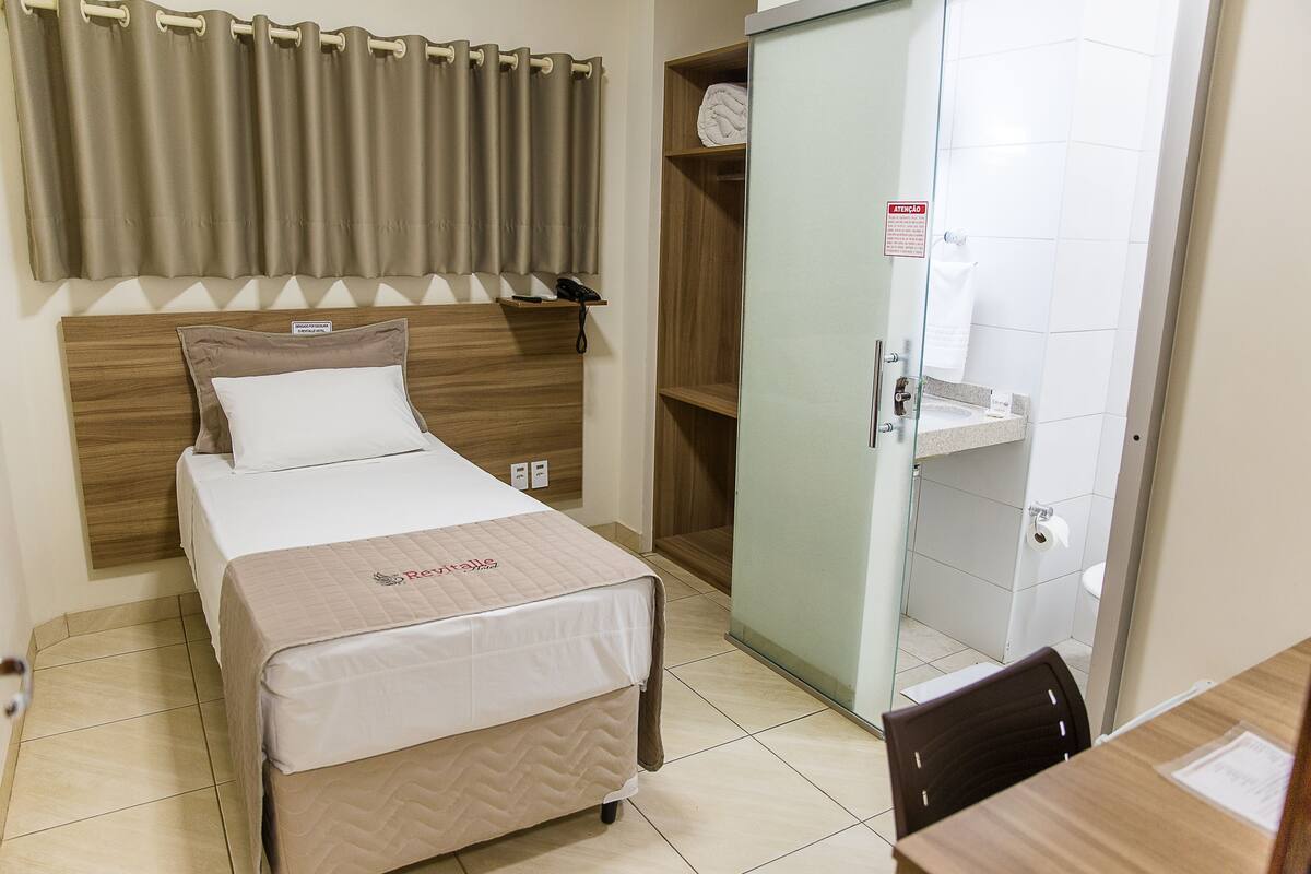 Ein Hotelzimmer mit einem Bett, einem hölzernen Kopfteil, einer Glastür zum Badezimmer und einem kleinen Tisch mit Stuhl.