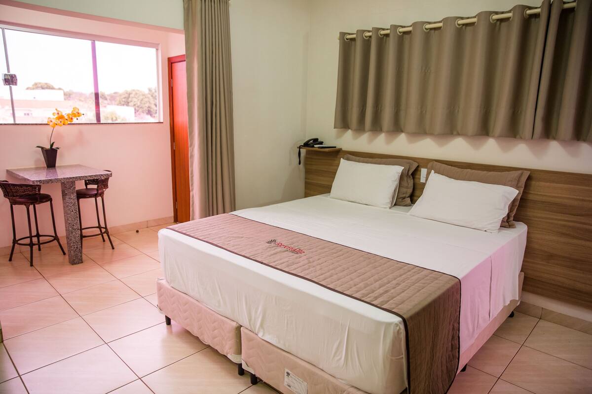 Ein Hotelzimmer mit einem Bett, einem Schreibtisch mit zwei Stühlen, einer Zimmerpflanze und einem Fenster mit Vorhängen.