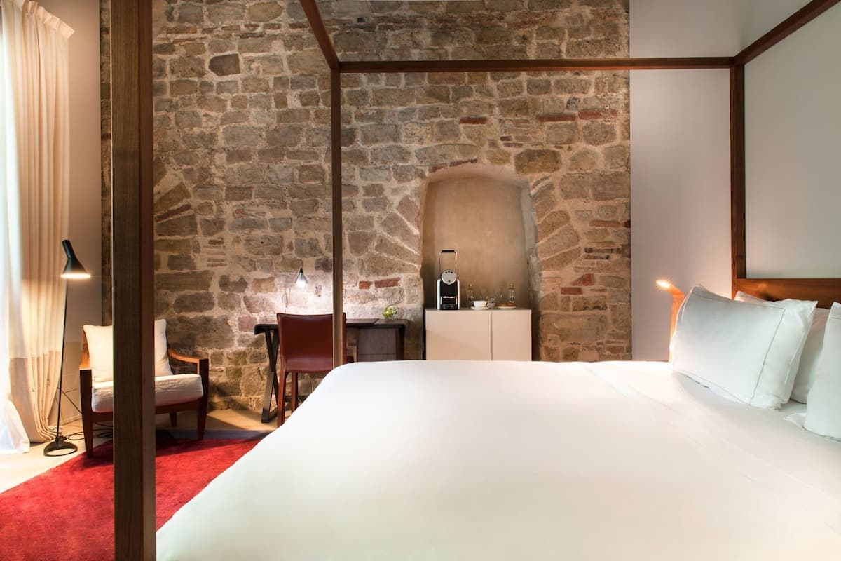 A hotel room with a stone wall, a bed with white linens, a desk with a lamp, and a chair.