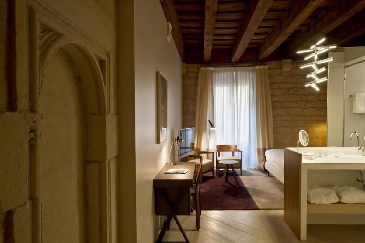 A bedroom with a wooden ceiling, a desk with a chair, a bed, and a bathroom with a mirror and sink.