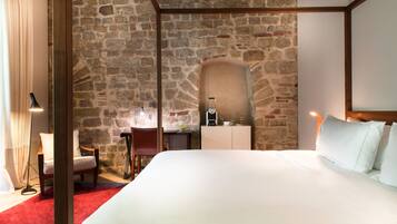 Un dormitorio con pared de piedra, una cama con ropa de cama blanca, cabecera de madera, un escritorio con silla y una mesita de noche con lámpara.