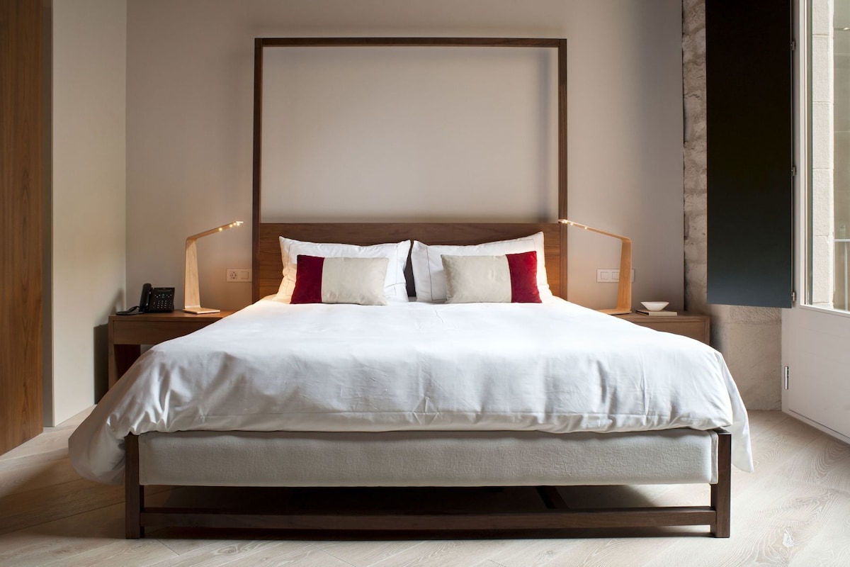 Una cama bien hecha con cabecera de madera, una mesita de noche con lámpara y vistas al exterior por una ventana.