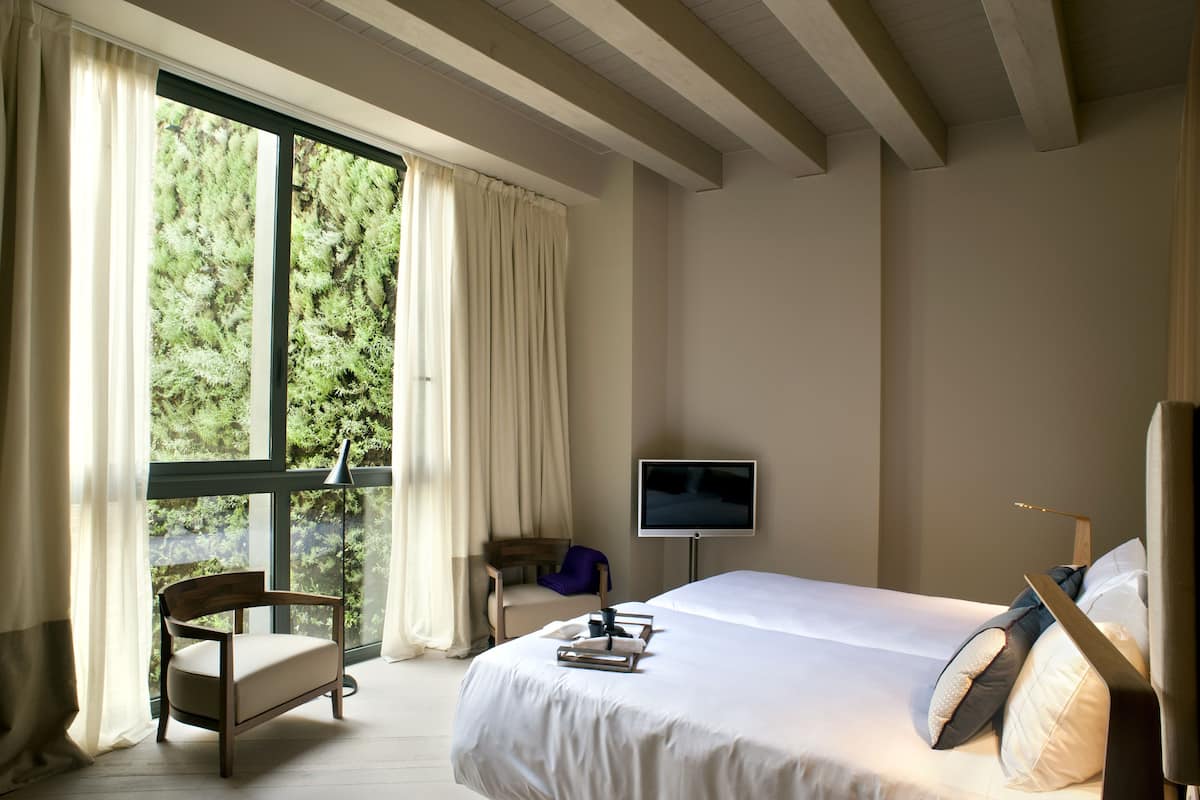 A bedroom with a large window, a bed, a chair, a flat-screen TV, and a bedside table.