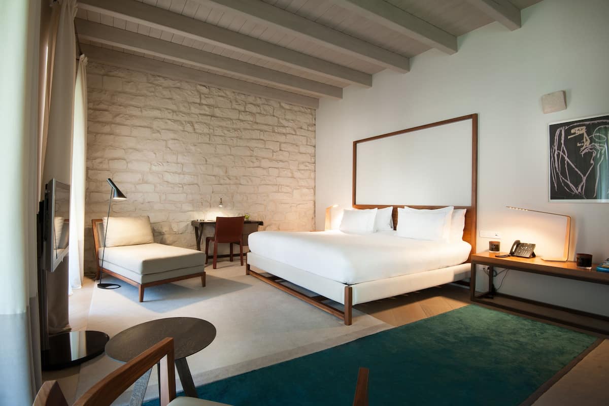 A bedroom with a bed, a chair, a desk, a lamp, and a painting on the wall.