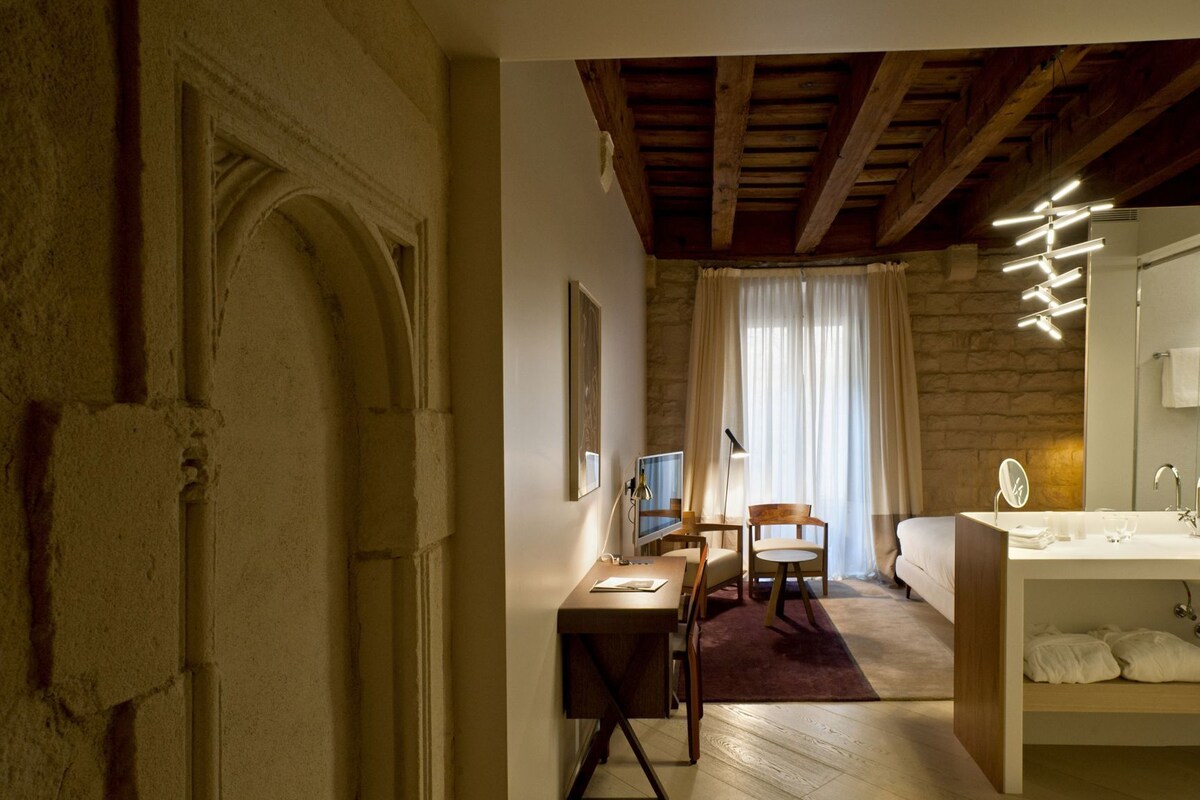 Una habitación con techo de madera, pared de piedra, cama, escritorio con silla, televisor y baño con espejo y lavamanos.