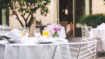 Mesa dispuesta para comer al aire libre con mantel blanco, un tazón, un vaso de agua y un vaso de jugo de naranja, además de un jarrón con flores.