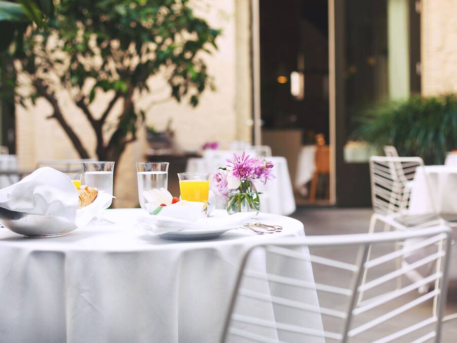 Mesa dispuesta para comer al aire libre con mantel blanco, un tazón, un vaso de agua y un vaso de jugo de naranja, además de un jarrón con flores.