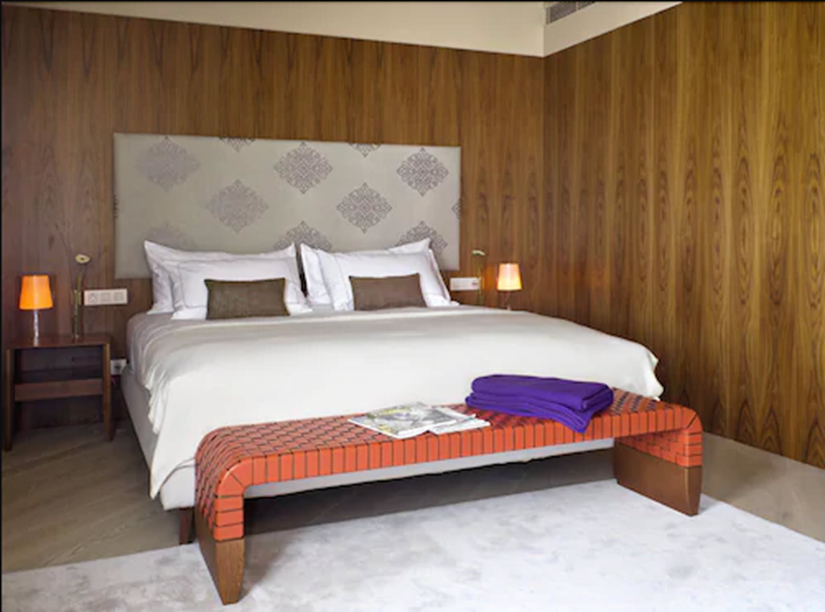 Une chambre avec un grand lit, des murs recouverts de panneaux en bois, deux tables de chevet avec des lampes et un banc au pied du lit.