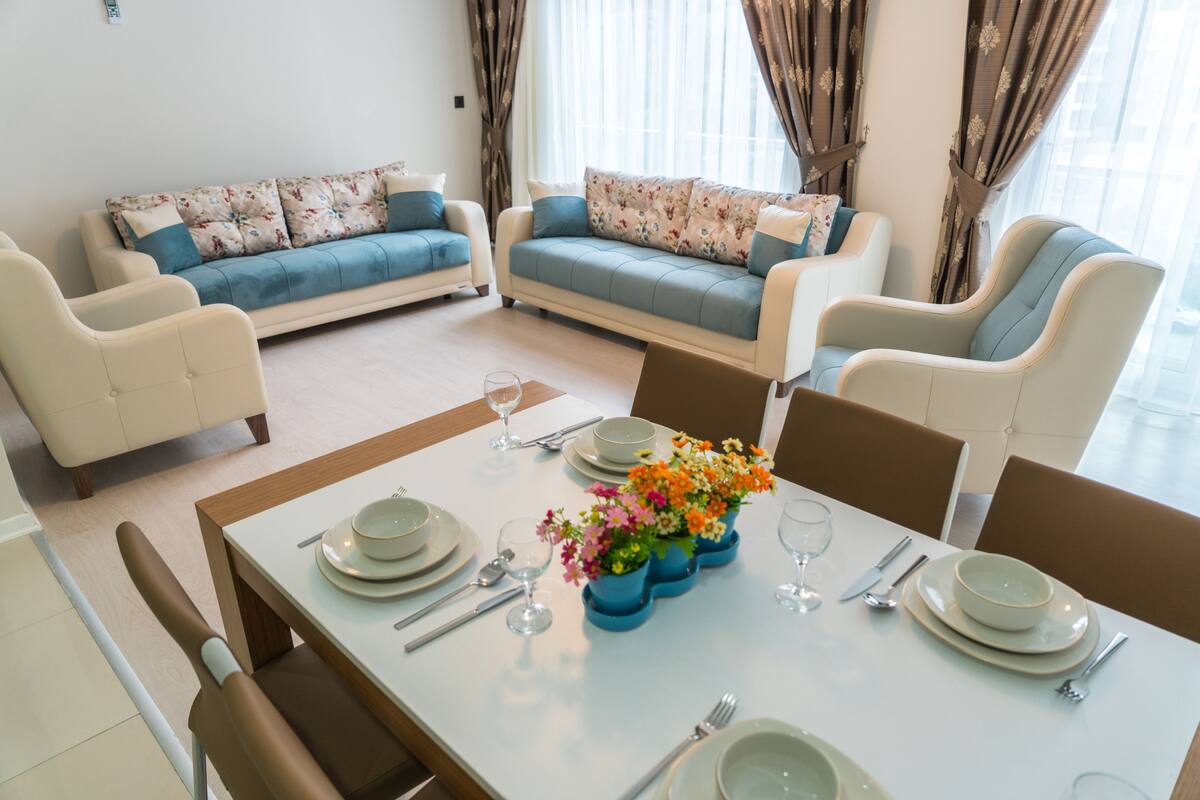 Un comedor con una mesa dispuesta para cuatro, con platos, tazones y cubiertos blancos. Alrededor de la mesa hay sillas azules y un arreglo floral en un jarrón.