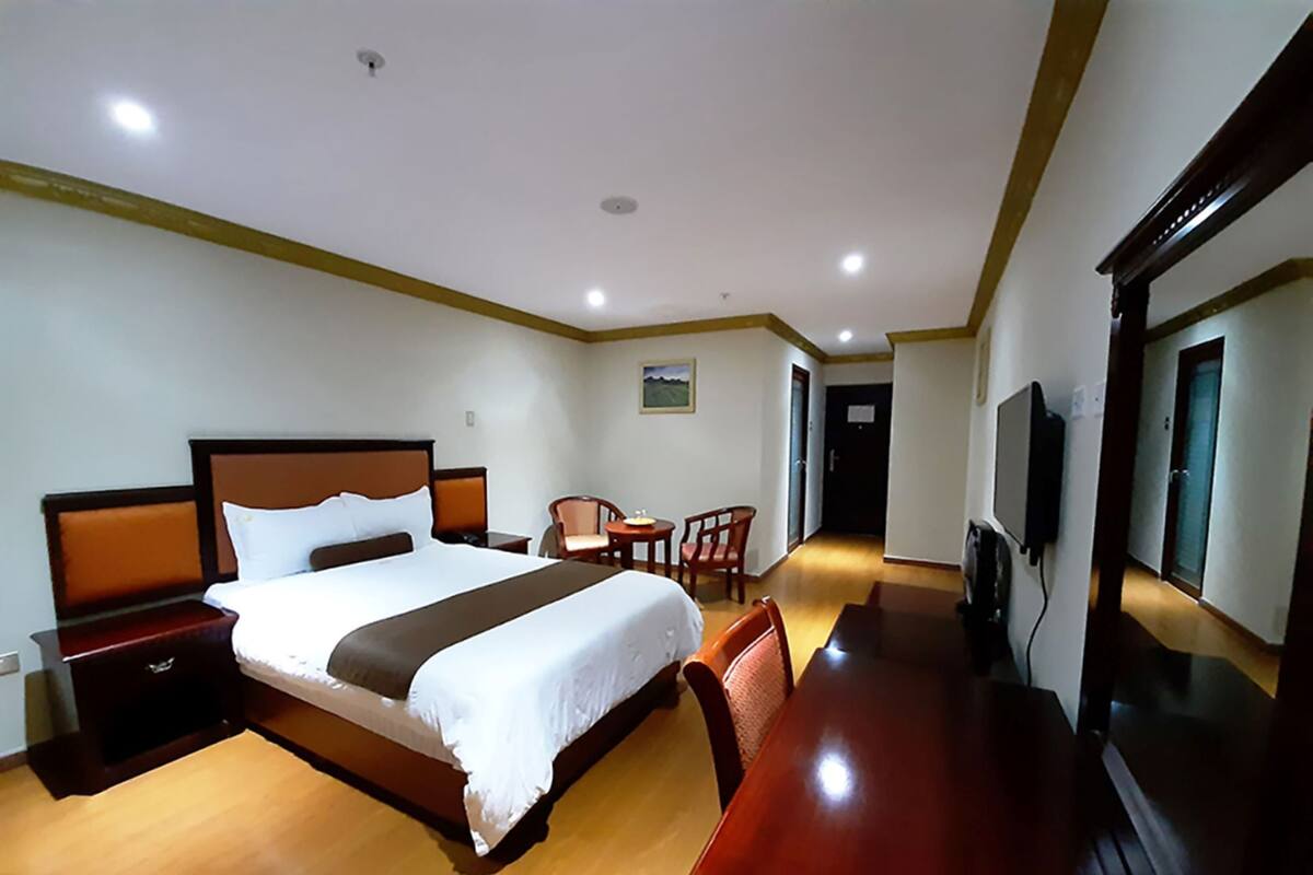 A hotel room with a large bed, a wooden desk, a chair, a television, and a door leading to another room.