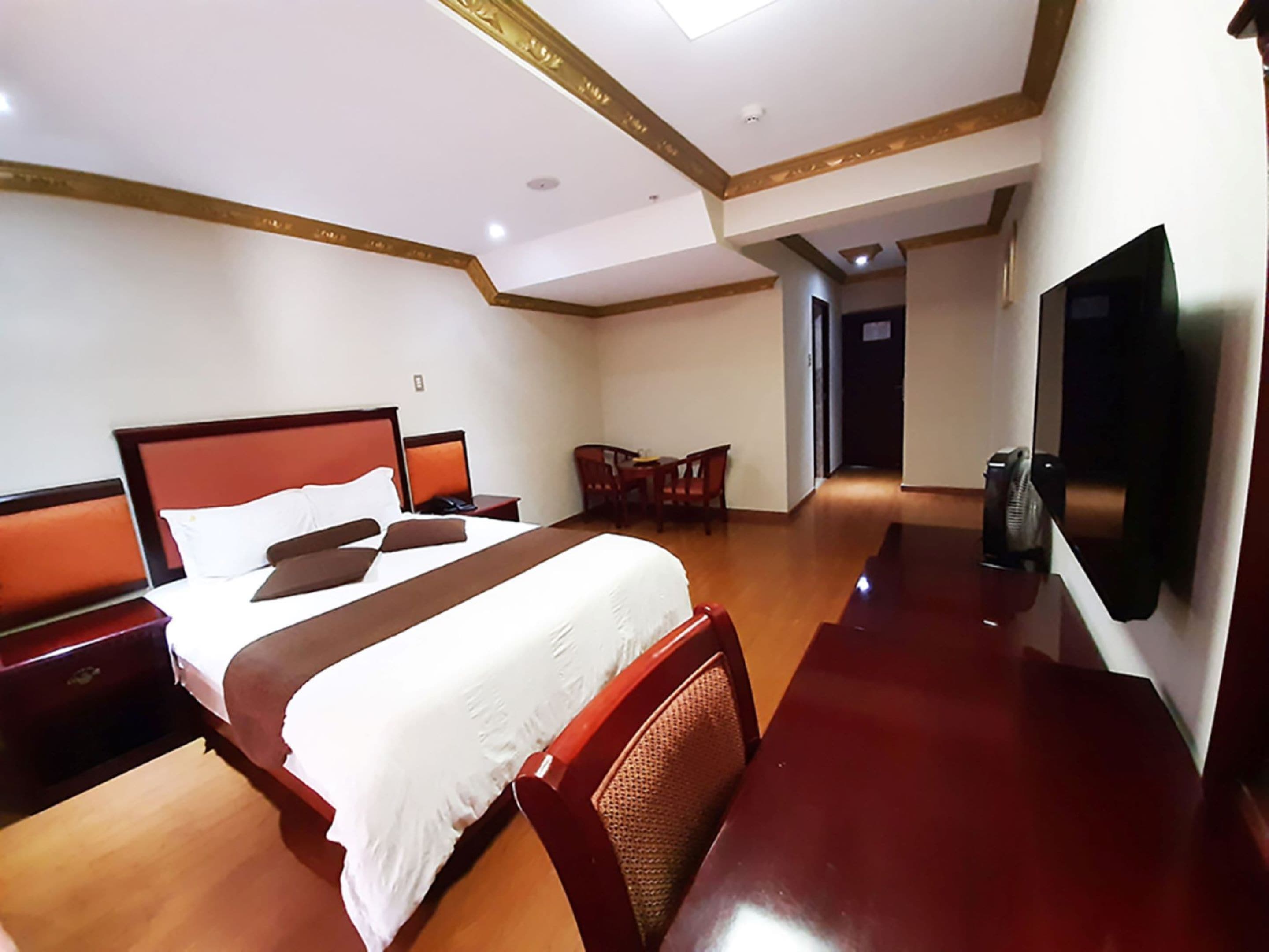 A hotel room with a large bed, a desk, a chair, a TV, and a door leading to another room.