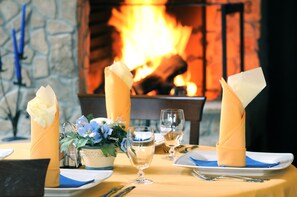 Mesa de comedor para dos personas, con servilletas blancas, un jarrón de flores y una chimenea al fondo.