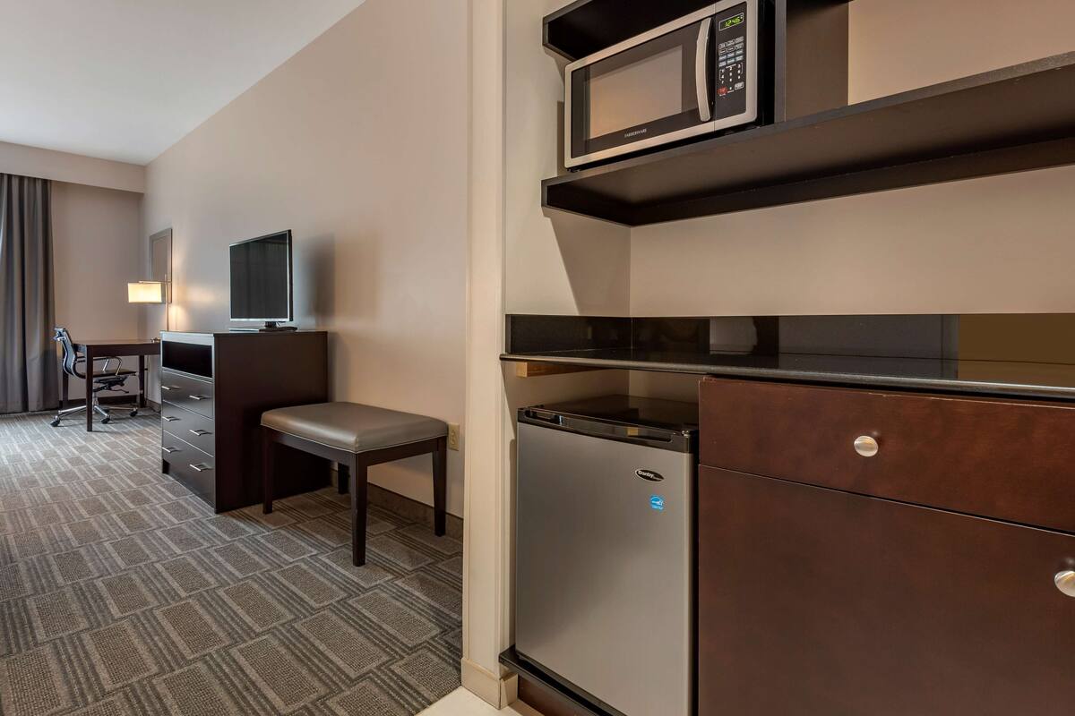A hotel room with a kitchenette including a microwave, refrigerator, and a small dining table with a chair.