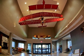 Een ruime lobby met een grote glazen ingang, zithoeken en een plafond met een rode hangende constructie.