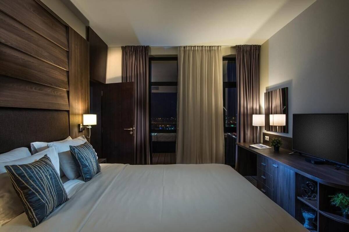 A hotel room with a large bed, two bedside lamps, a TV, and a view of the city at night.