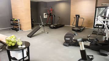 Un gimnasio con diversos equipos de ejercicio, una pequeña mesa con un bol de manzanas y vasos, y un espejo en la pared.