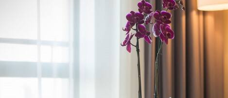 A hotel room with a wooden table, a bottle of wine, two wine glasses, a plate with food, a vase with purple flowers, and a pair of glasses on the table.