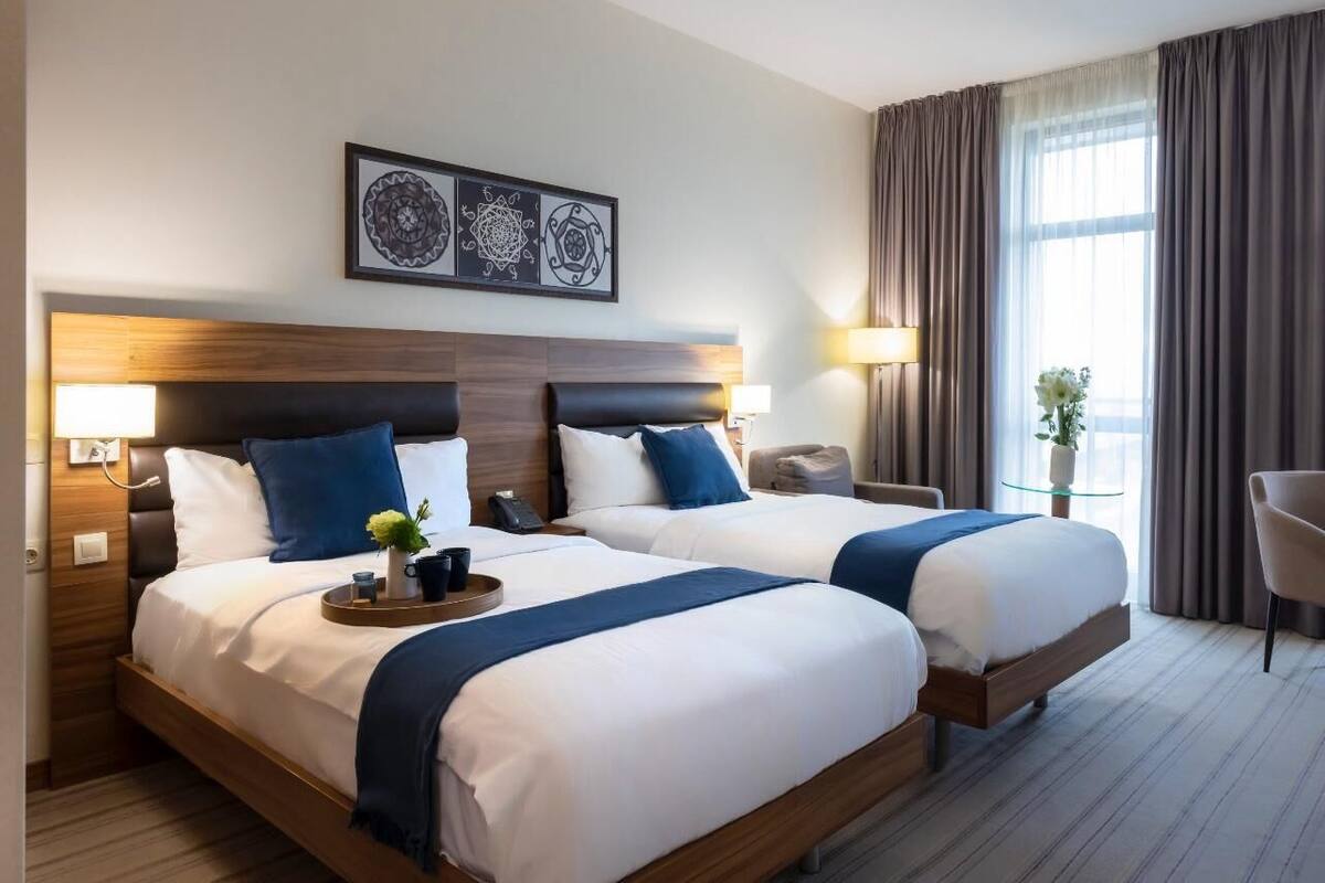 A hotel room with two beds, a wooden headboard, a nightstand with a lamp, a small table with a tray, and a chair.