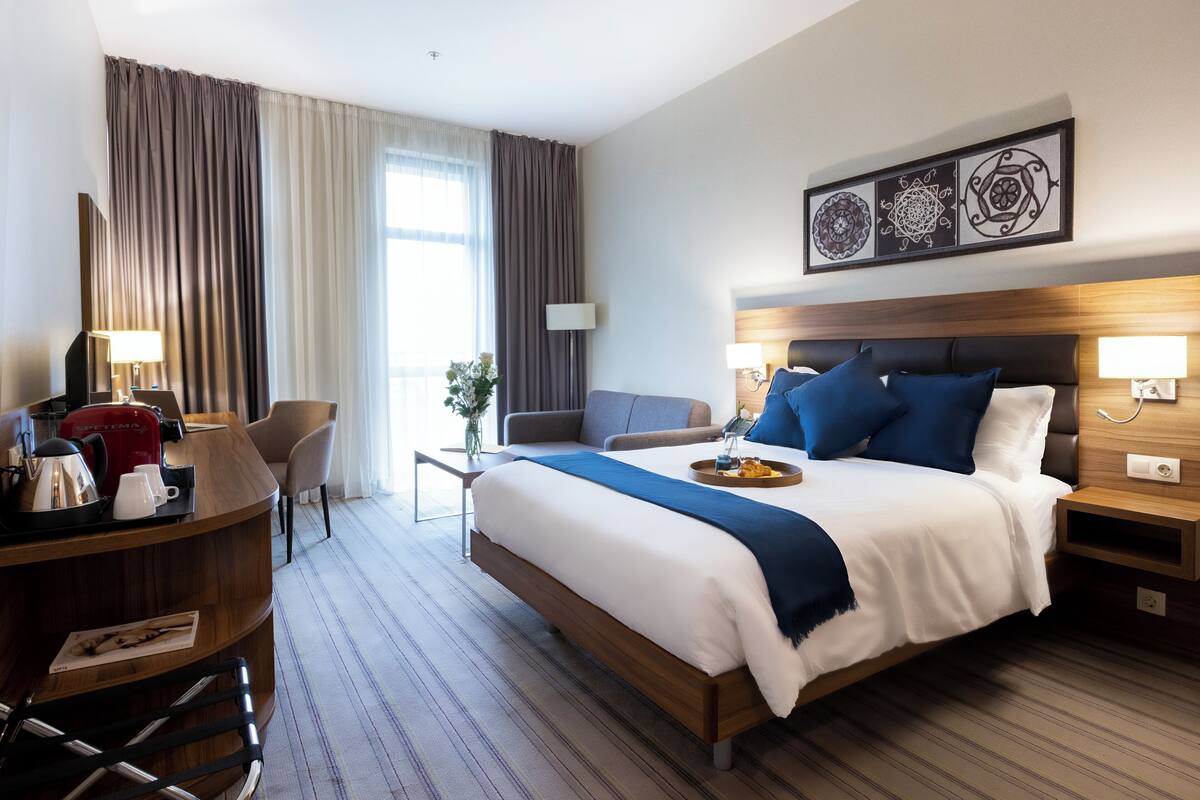 A hotel room with a large bed, a desk with a lamp, a chair, a sofa, and a coffee maker.