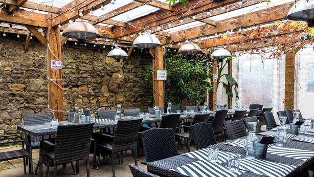 An outdoor patio with tables and chairs, stone walls, and hanging lights. - The First Hurdle Guest House (Chepstow)