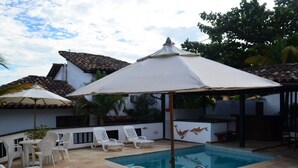 Un área de piscina con sillas reclinables blancas y sombrillas, un suelo de baldosas y una piscina.