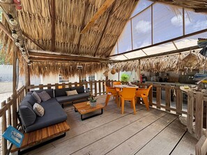 Een overdekt terras met rieten dak, houten meubilair en uitzicht op het omliggende landschap.