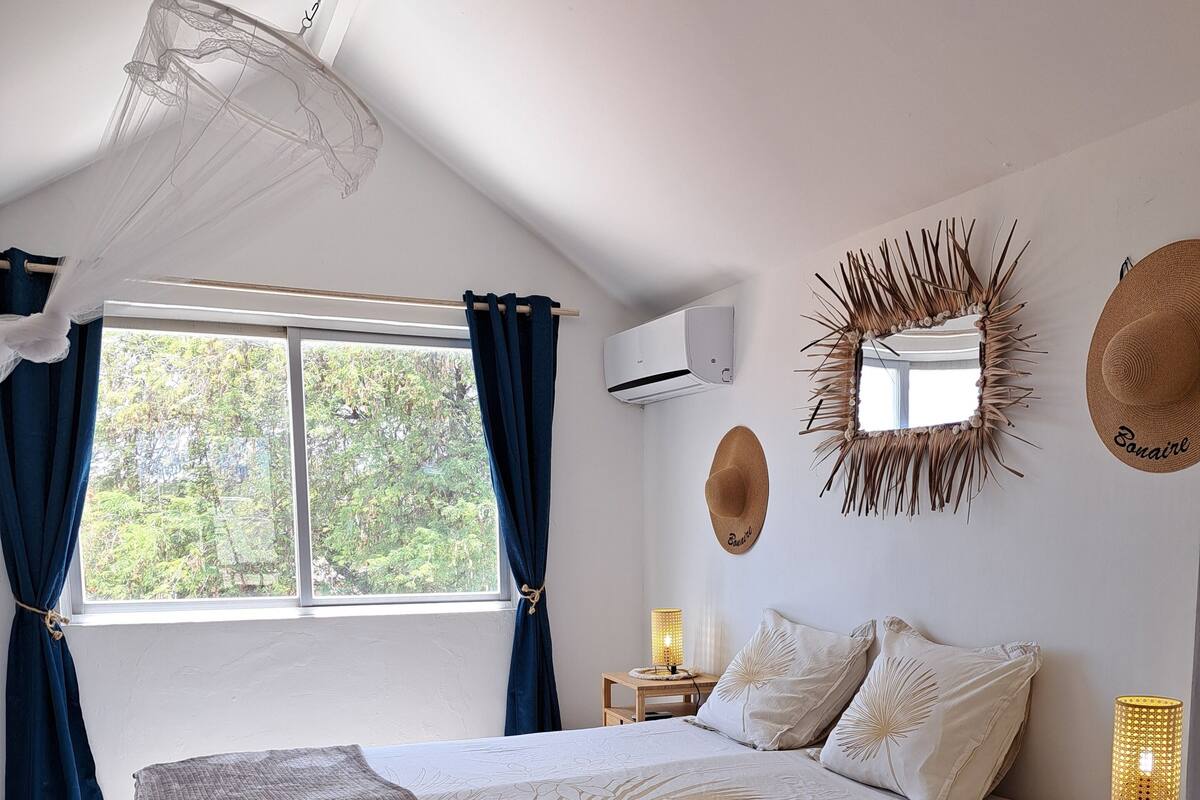 Une chambre à coucher avec un lit, une table de chevet avec une lampe, un miroir fixé au mur et une fenêtre avec des rideaux.