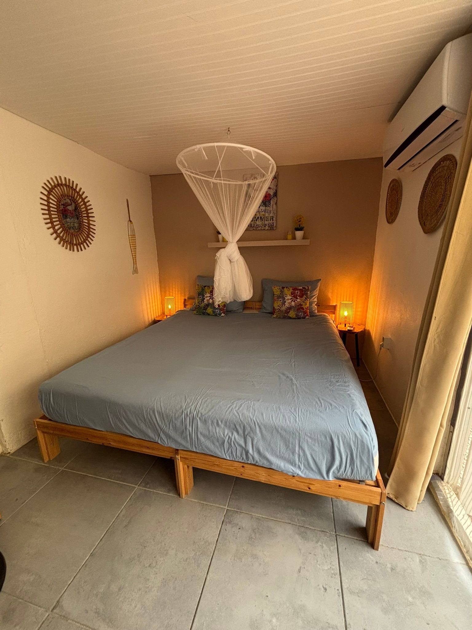 A bedroom with a bed, a hanging mosquito net, a wall decoration, a shelf with items, and an air conditioning unit.