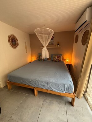 A bedroom with a bed, a hanging mosquito net, a wall decoration, a shelf with items, and an air conditioning unit.
