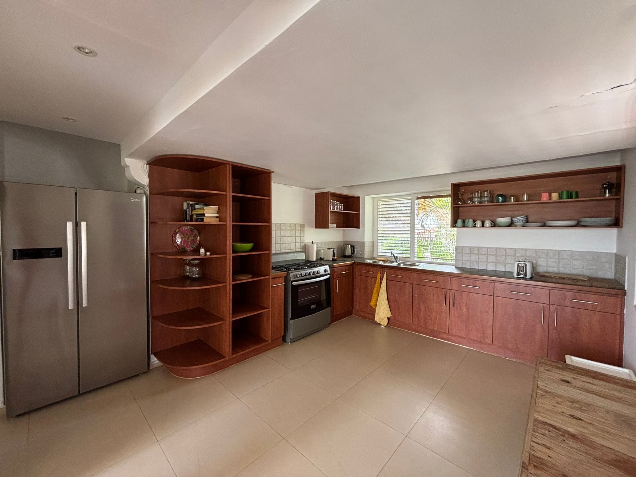 A kitchen with a built-in oven, a refrigerator, wooden shelves, and a window with blinds.