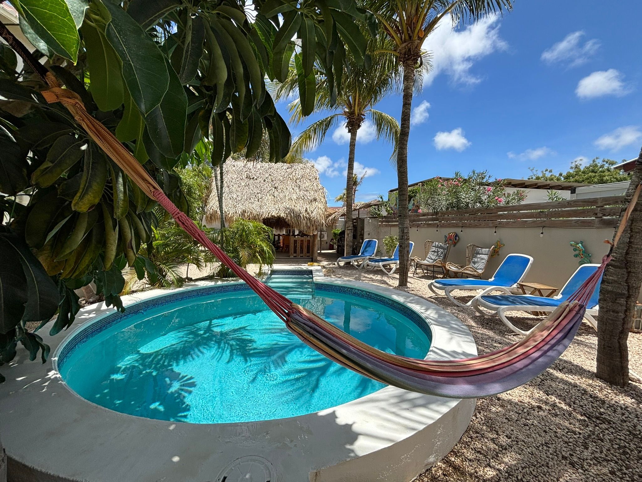 A swimming pool with a hammock, lounge chairs, palm trees, and a thatched-roof structure.