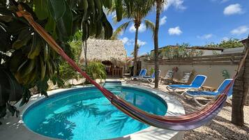A swimming pool with a hammock, lounge chairs, palm trees, and a thatched-roof structure.