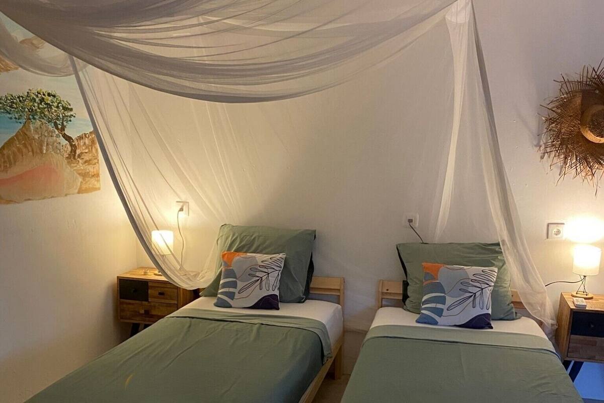 Une chambre avec deux lits en bois, chacun agrémenté de draps verts et d’oreillers décoratifs. Un baldaquin recouvre les lits, et il y a des tables de chevet avec des lampes.