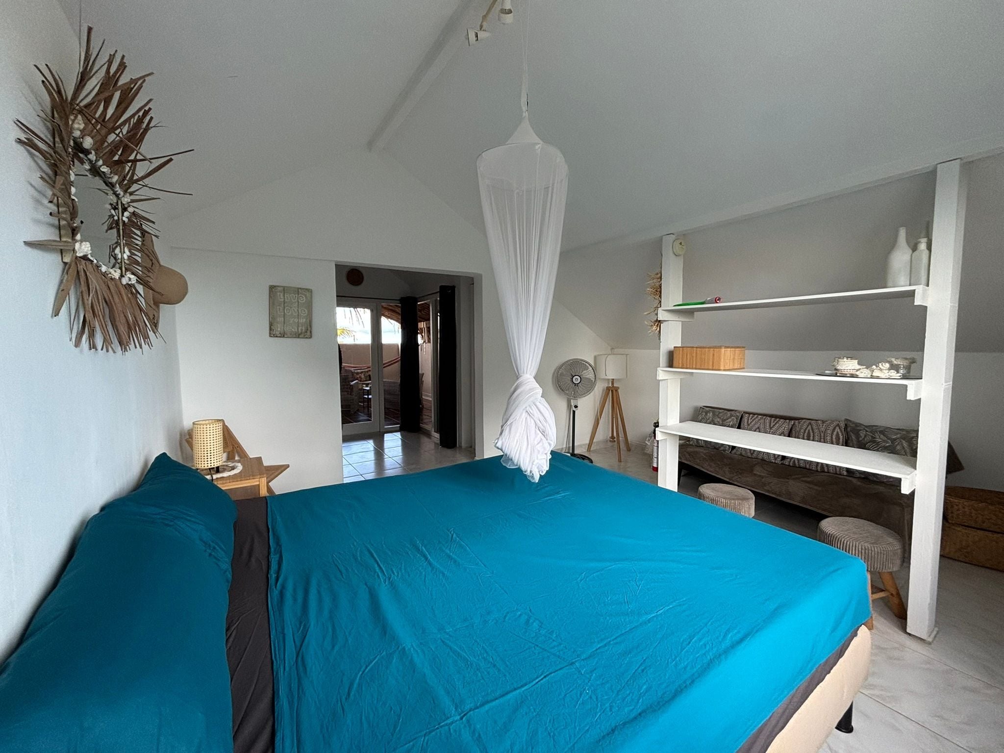 A bedroom with a bed, a bedside table, a lamp, a shelf with decorative items, and a doorway leading to another room.