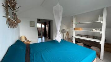 A bedroom with a bed, a bedside table, a lamp, a shelf with decorative items, and a doorway leading to another room.