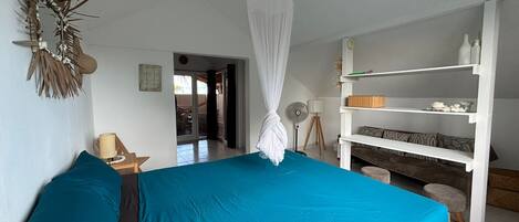 Un dormitorio con cama, mesita de noche, lámpara, estante con adornos y una puerta que da a otra habitación.