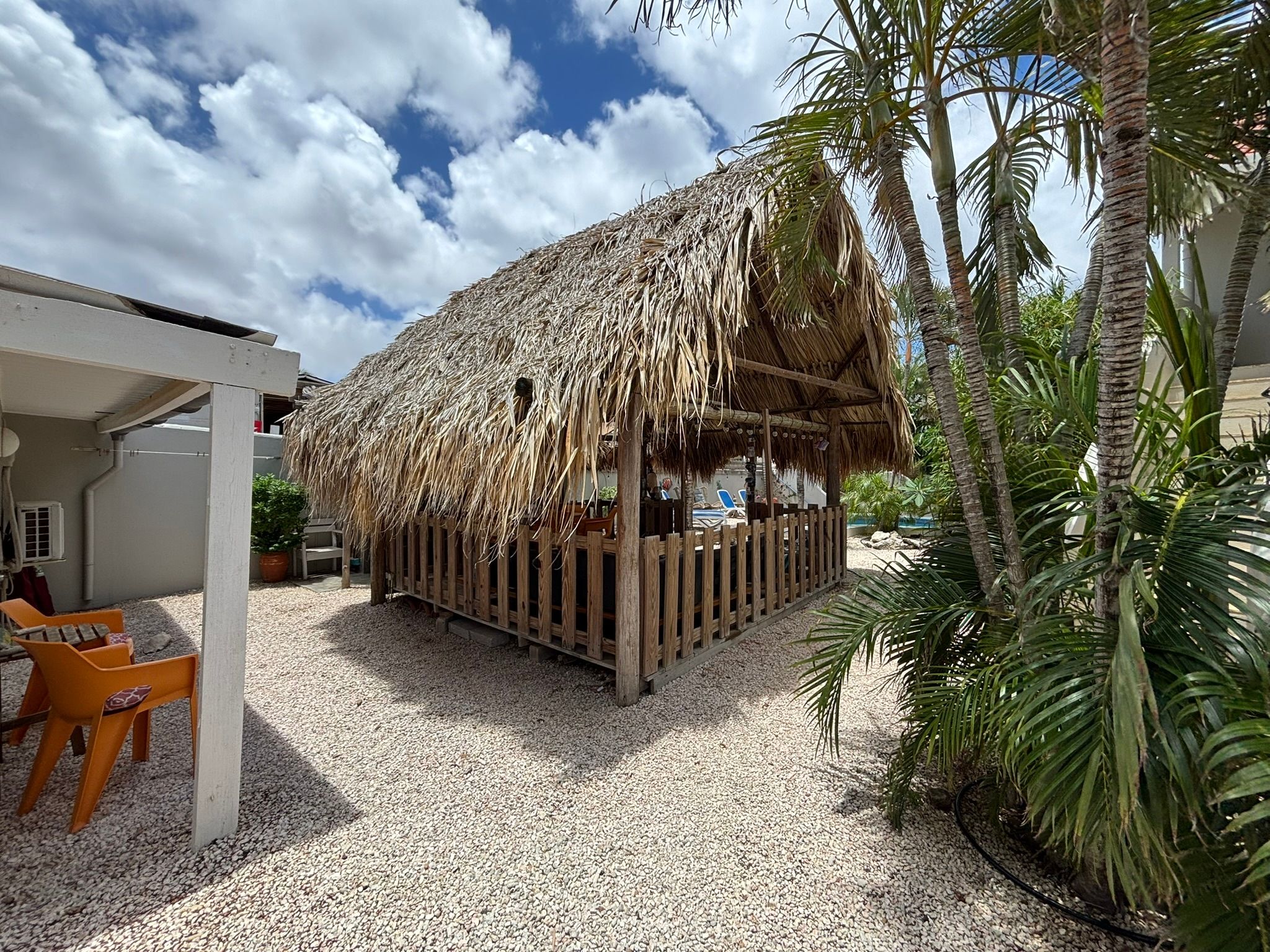 A thatched-roof structure with a wooden fence, surrounded by gravel and palm trees.