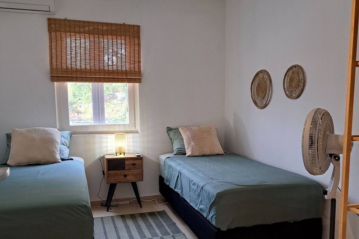 Une petite chambre bien éclairée, avec deux lits simples, une table de chevet avec une lampe, une fenêtre avec un store en bambou et un ventilateur sur pied.