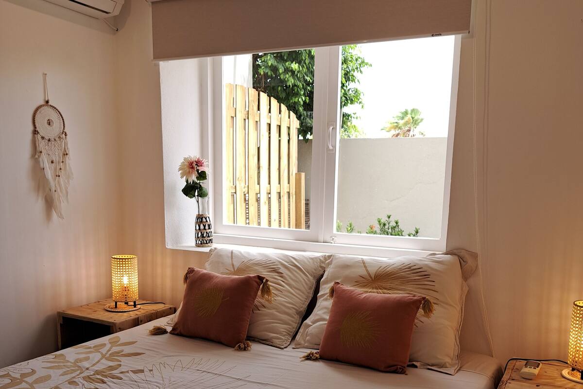 Une chambre à coucher avec un grand lit, deux tables de chevet avec des lampes, une fenêtre donnant sur une clôture en bois et de la verdure, et un climatiseur fixé au mur.